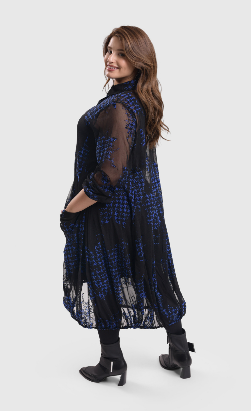 Left side/back full body view of a woman wearing the alembika floral check wonderful dress. This long sleeved dress is black mesh/sheer with blue and black houndstooth/floral printed pieces of fabric all over it.