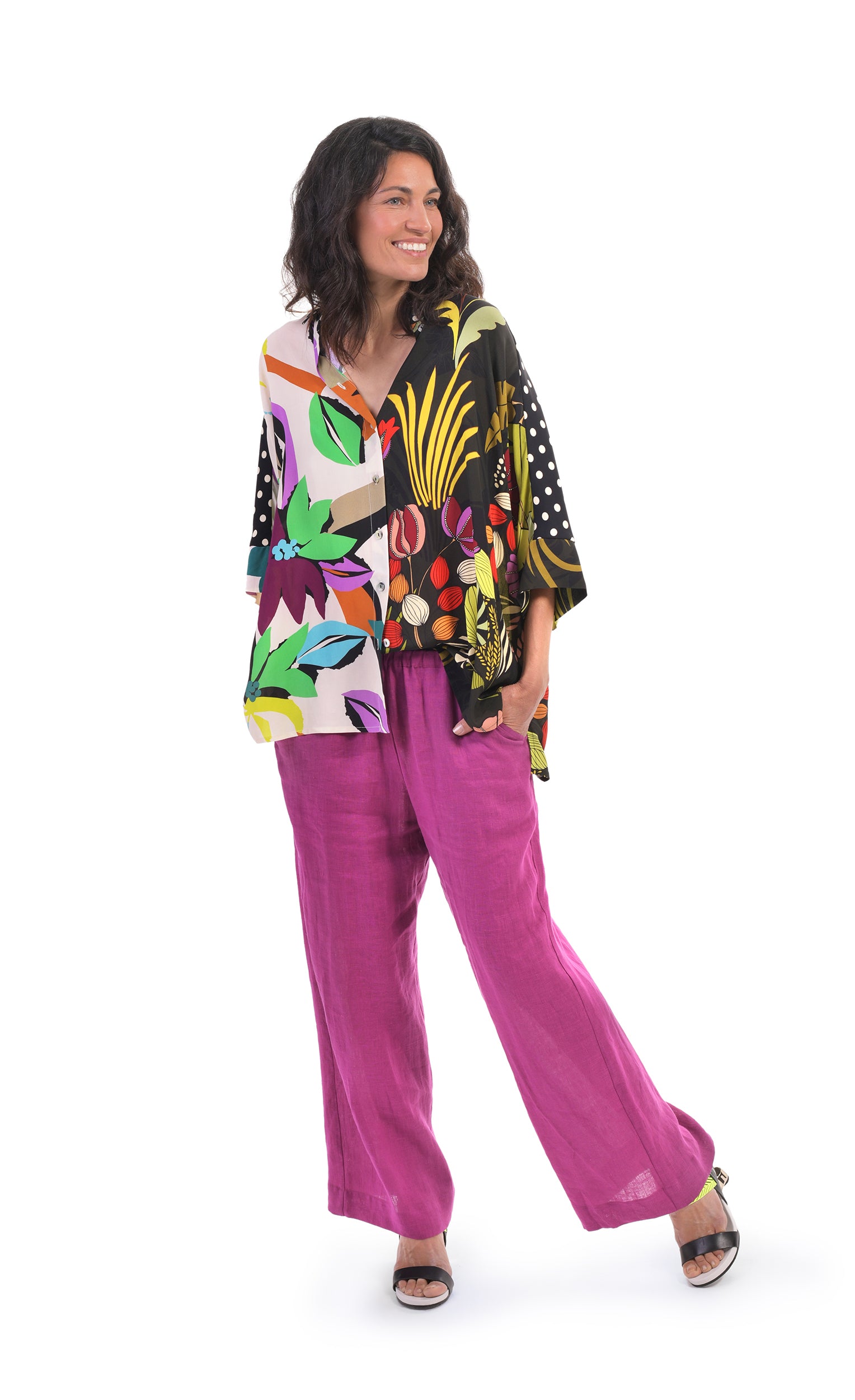 Front full body view of the alembika floral mix print campshirt. This shirt  has a light colored jungle print on the right side and a dark colored jungle print on the left side. The shirt has a button down front, dotted sleeves, a relaxed fit, and drop shoulders. On the bottom she is wearing pink pants.