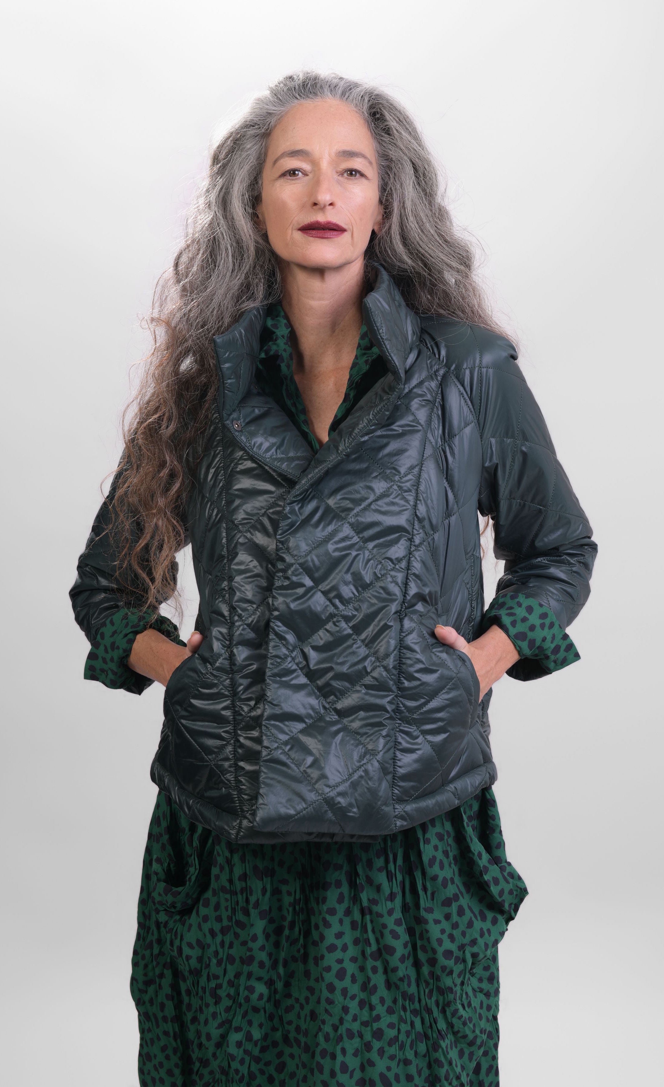 Front top half view of a woman wearing the alembika forest green jacket over the alembika cheetah print dress in pine green. This jacket is a puffer jacket with a tall stand collar, long sleeves, and a hidden button up front.