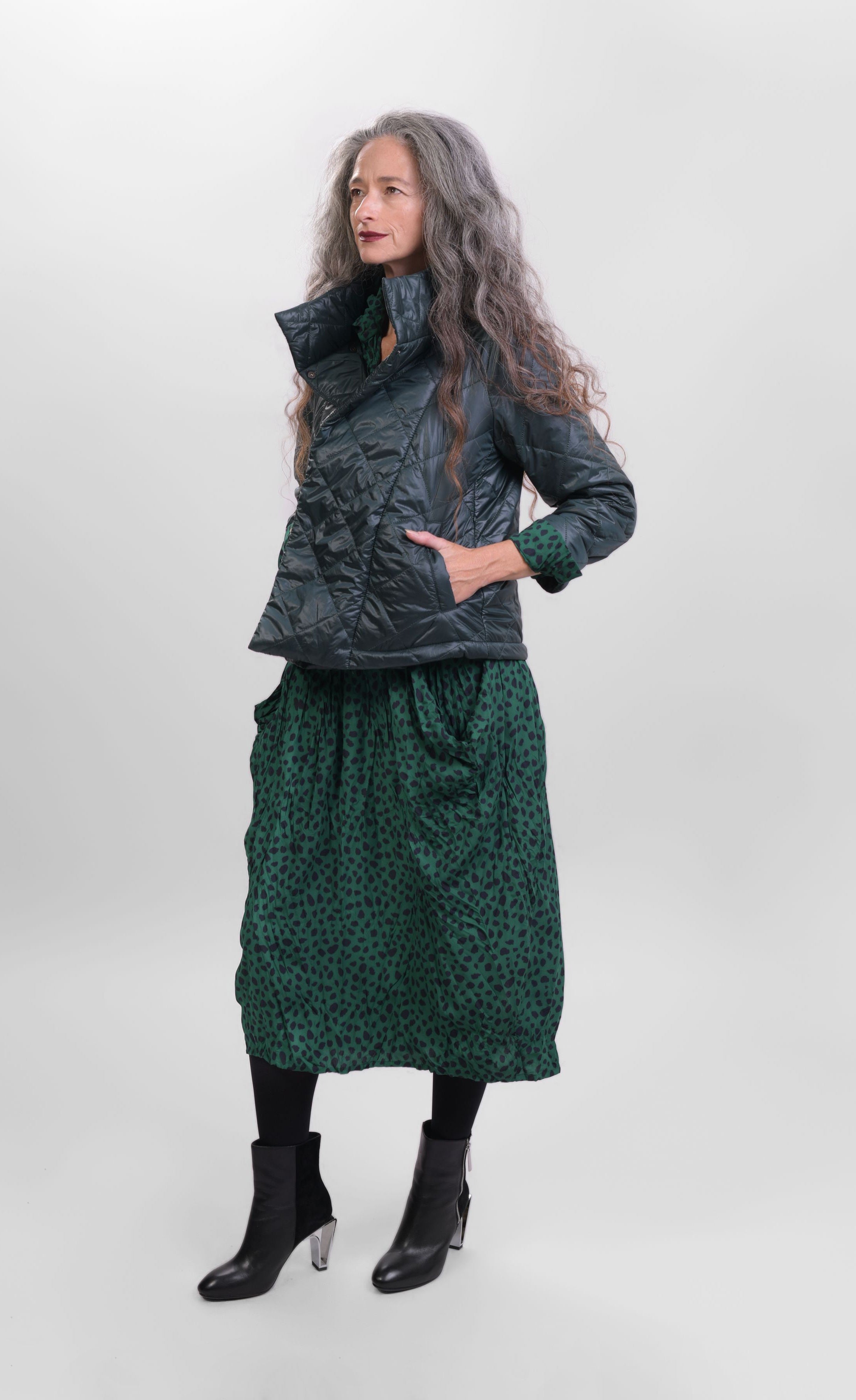 Front full body view of a woman wearing the alembika forest green jacket over the alembika cheetah print dress in pine green. This jacket is a puffer jacket with a tall stand collar, long sleeves, and a hidden button up front.