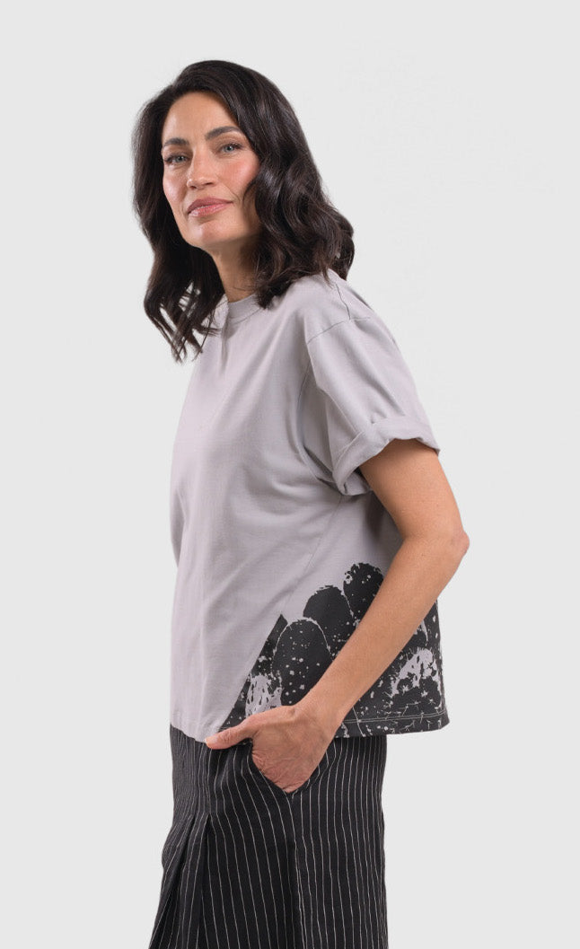 Left side top half view of a woman wearing the alembika grey cactus tee. This top is solid grey with a black stamped cactus print on the bottom of the left side. The top has short sleeves that are rolled up and a boxy silhouette.