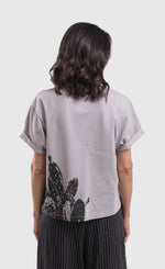 Load image into Gallery viewer, Back  top half view of a woman wearing the alembika grey cactus tee. This top is solid grey with a black stamped cactus print on the bottom of the left side. The top has short sleeves that are rolled up and a boxy silhouette.
