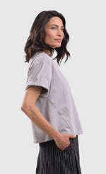 Load image into Gallery viewer, Right side top half view of a woman wearing the alembika grey cactus tee. This top is solid grey with a black stamped cactus print on the bottom of the left side. The top has short sleeves that are rolled up and a boxy silhouette.

