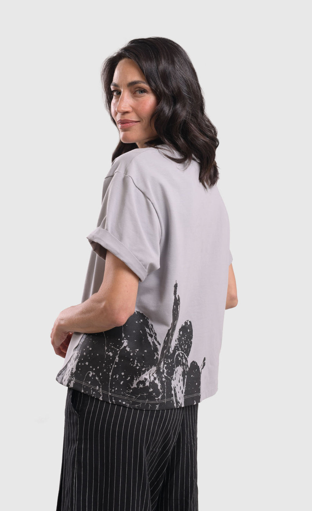 Back side top half view of a woman wearing the alembika grey cactus tee. This top is solid grey with a black stamped cactus print on the bottom of the left side. The top has short sleeves that are rolled up and a boxy silhouette.