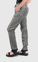 Load image into Gallery viewer, left side bottom half view of a woman wearing the alembika iconic stretch jeans in a pinstripe black and white print. This pant has a drawstring waistband with a tie, two front pockets, two back pockets, and a relaxed slim leg.
