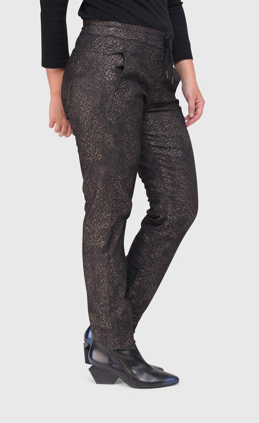 Bottom half right side view of a woman wearing the alembika iconic stretch pant in the color sepia. The pant is dark brown/black with a small metallic-like animal print. It also has a drawstring and tie waistband.