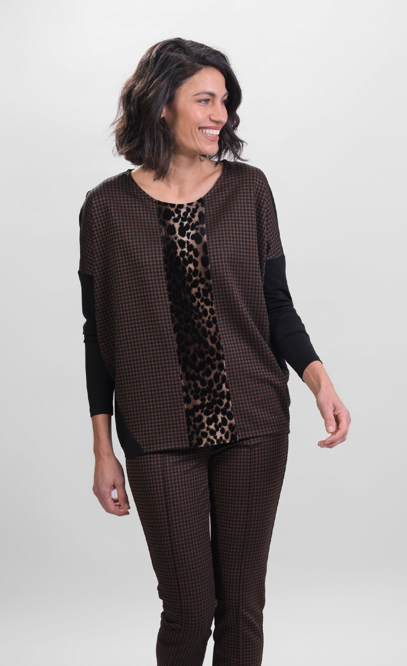 Front top half view of a woman wearing brown pants and the alembika leopard panel top. This top has a round neck, solid black drop shoulder dolman sleeves, a brown body, and a leopard print panel running down the center of the front.