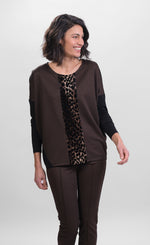 Load image into Gallery viewer, Front top half view of a woman wearing brown pants and the alembika leopard panel top. This top has a round neck, solid black drop shoulder dolman sleeves, a brown body, and a leopard print panel running down the center of the front.
