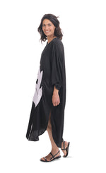 Load image into Gallery viewer, Left side full body view of a woman wearing the alembika lotus caftan dress in black. This dress sits below the knees. It has 3/4 length sleeves, side slits, and a large white lotus flower on the front.
