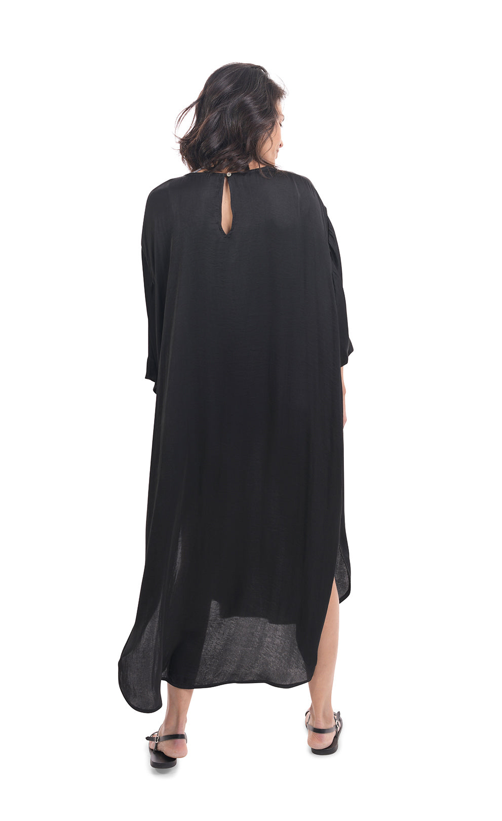 Back full body view of a woman wearing the alembika lotus caftan dress in black. This dress sits below the knees. It has 3/4 length sleeves, side slits, and keyhole back