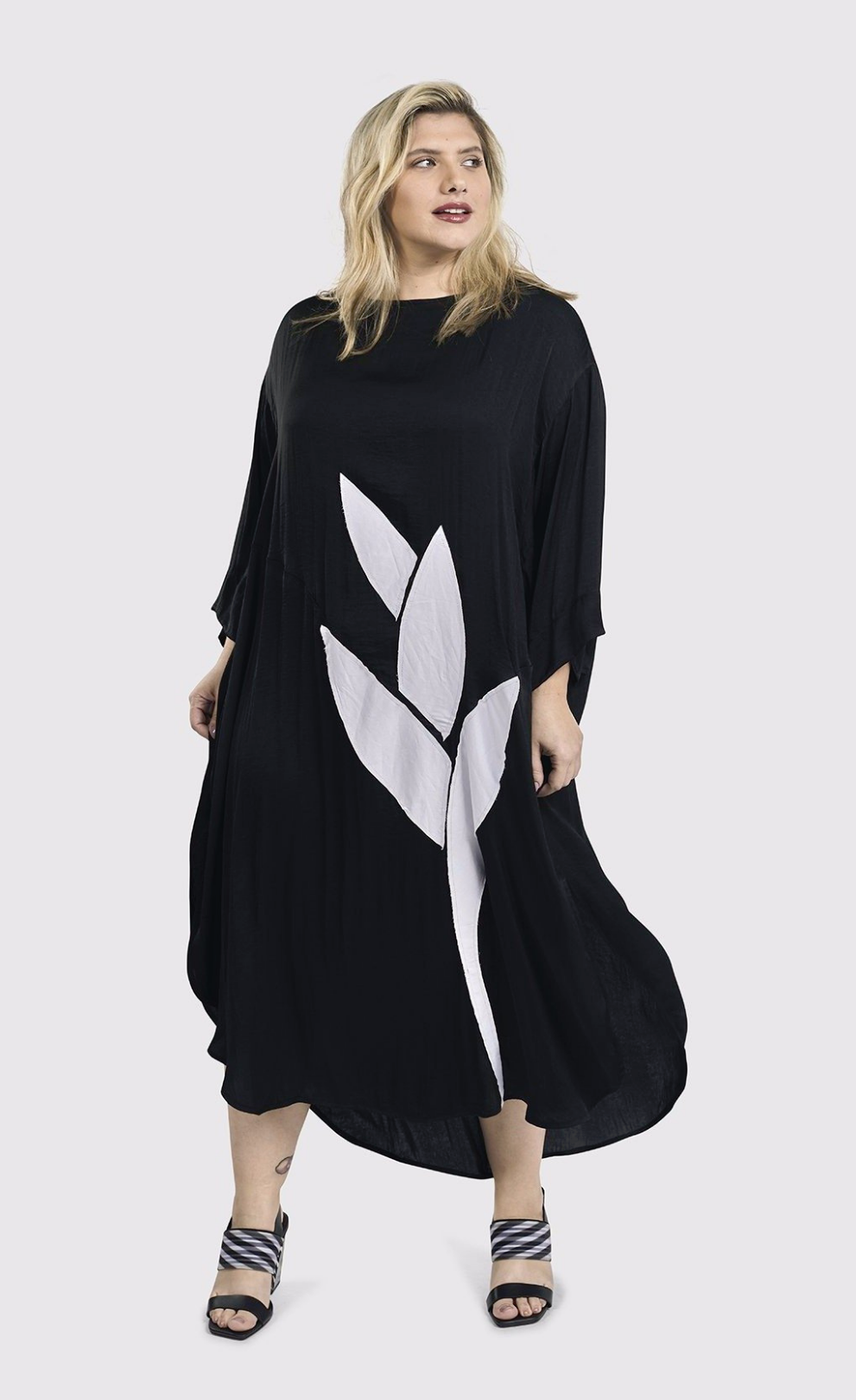 Front full body view of a woman wearing the alembika lotus caftan dress in black. This dress sits below the knees. It has 3/4 length sleeves, side slits, and a large white lotus flower on the front.