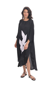 Front full body view of a woman wearing the alembika lotus caftan dress in black. This dress sits below the knees. It has 3/4 length sleeves, side slits, and a large white lotus flower on the front.
