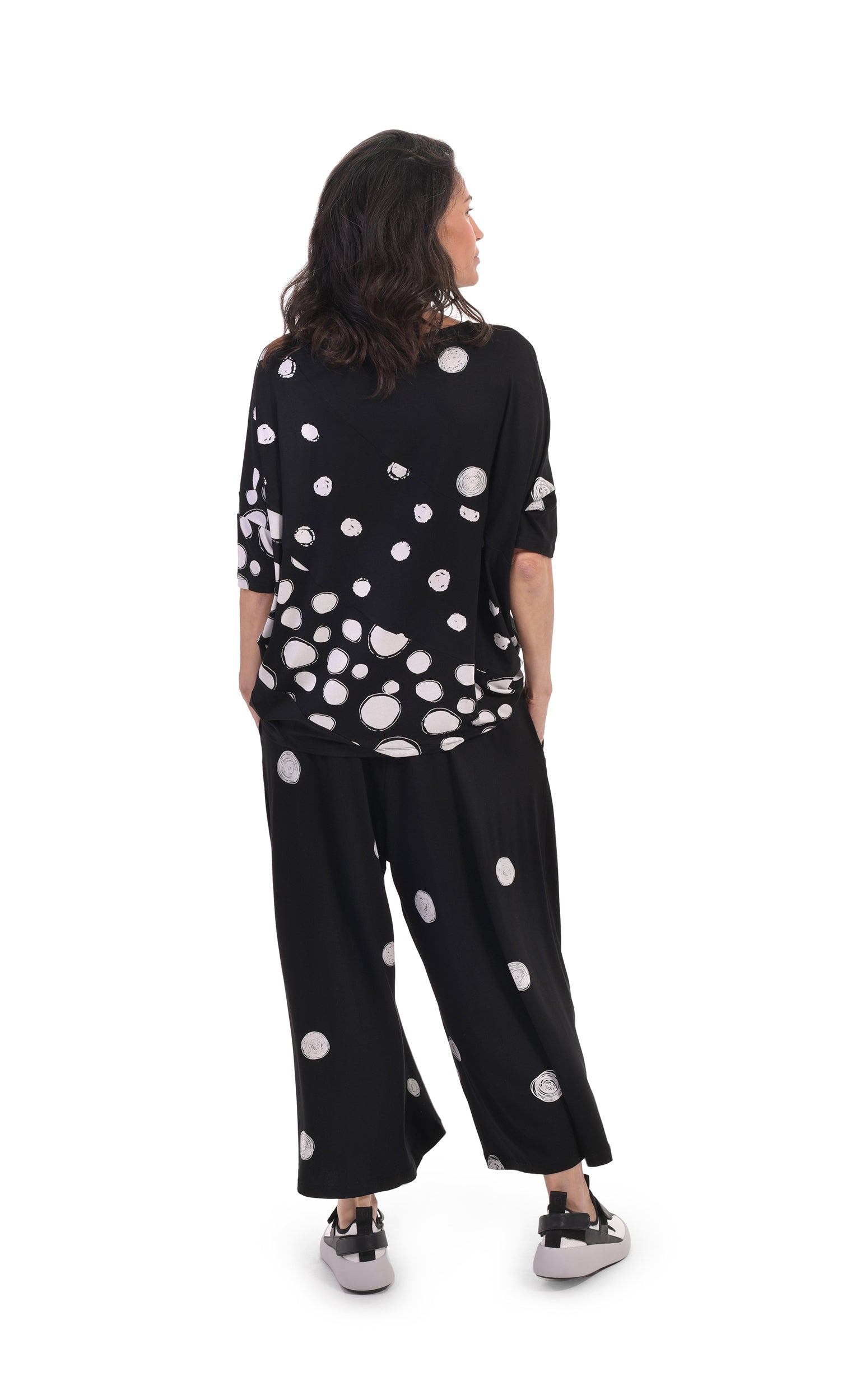Back, full body view of a woman wearing the alembika multi spotted lia jersey top. This top is black with different types of white spots all over it. The sleeves are 3/4 length and the shirt has an oversized fit. On the bottom she is wearing full leg black pants with white dots.