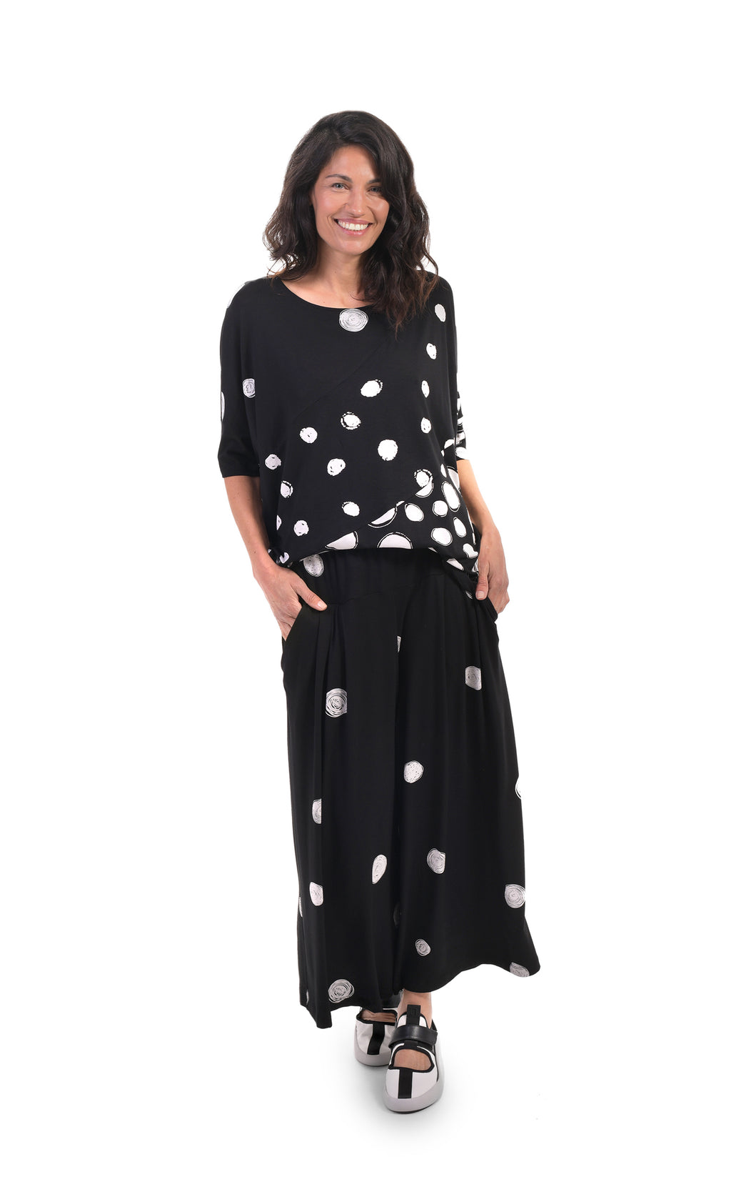 Front full body view of a woman wearing the alembika multi spotted lia jersey top. This top is black with different types of white spots all over it. The sleeves are 3/4 length and the shirt has an oversized fit.
