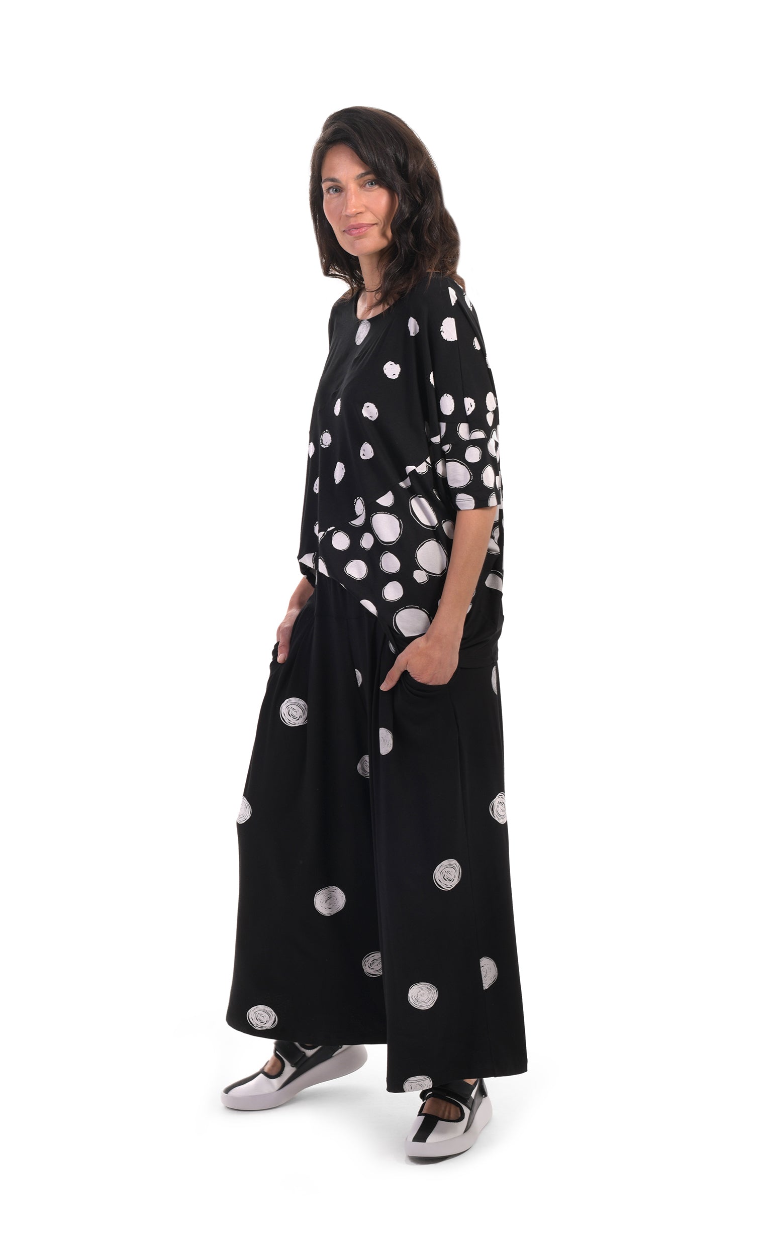 Front, left sided, full body view of a woman wearing the alembika multi spotted lia jersey top. This top is black with different types of white spots all over it. The sleeves are 3/4 length and the shirt has an oversized fit. On the bottom she is wearing full leg black pants with white dots.