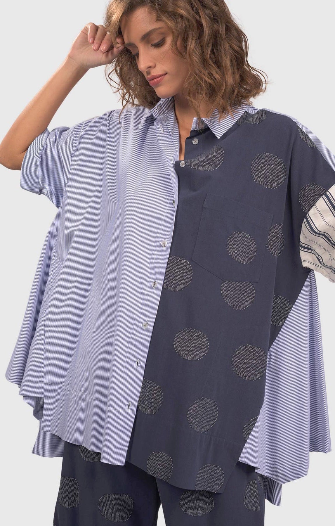 Front top half view of a woman wearing blue pants with green dots and the alembika navy short sleeve shirt. This shirt has blue and white stripes on the front right side and blue with green dots on the left side.