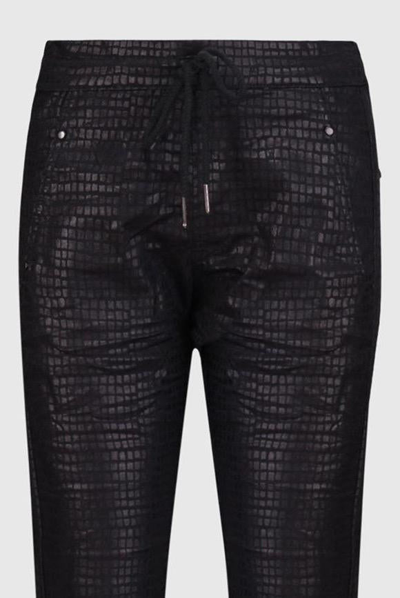 Front close up view of the Alembika Onyx Pant. This pant is black with a shiny snakeskin print on it. It has a drawstring waistband, slim legs, and two slanted side pockets.