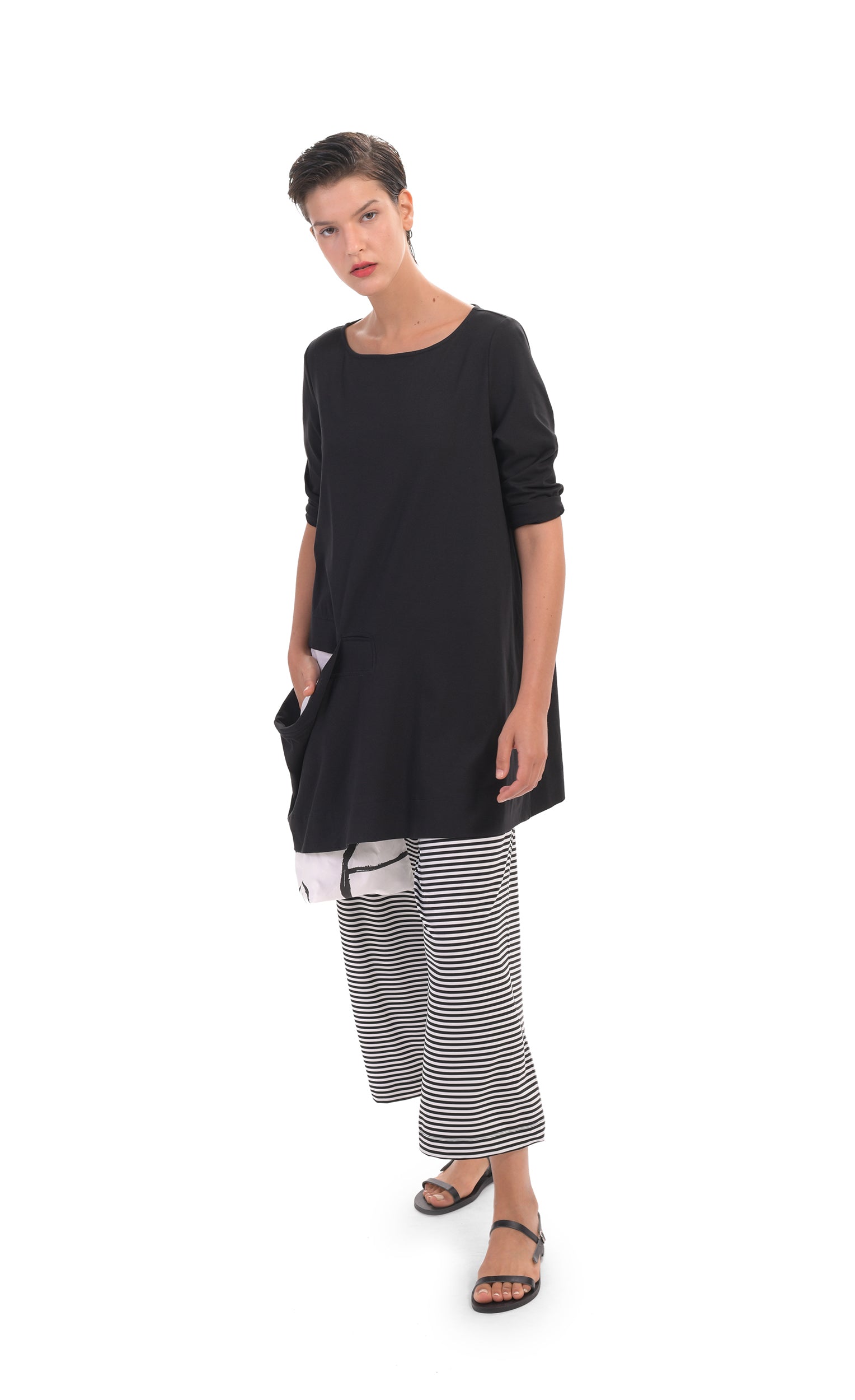 Front full body view of a woman wearing the alembika peek-a-boo pocket tunic with black and white striped pants. The top has 3/4 length sleeves. It is black with a draped open pocket. The pocket is lined with a white and black circle/capsule like print that hangs down below the hem.