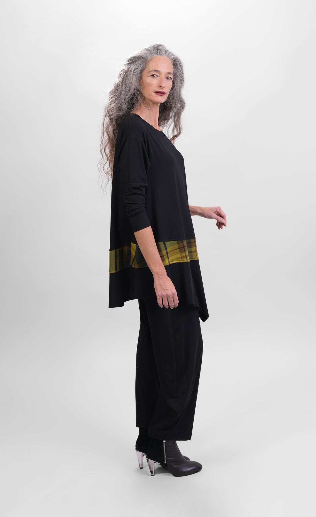 Left side full body view of a woman wearing black pants and the alembika plaid asymmetry top. This top is black with long sleeves and a horizontal green and yellow plaid panel running across the bottom of the top. The from hem is asymmetrical.