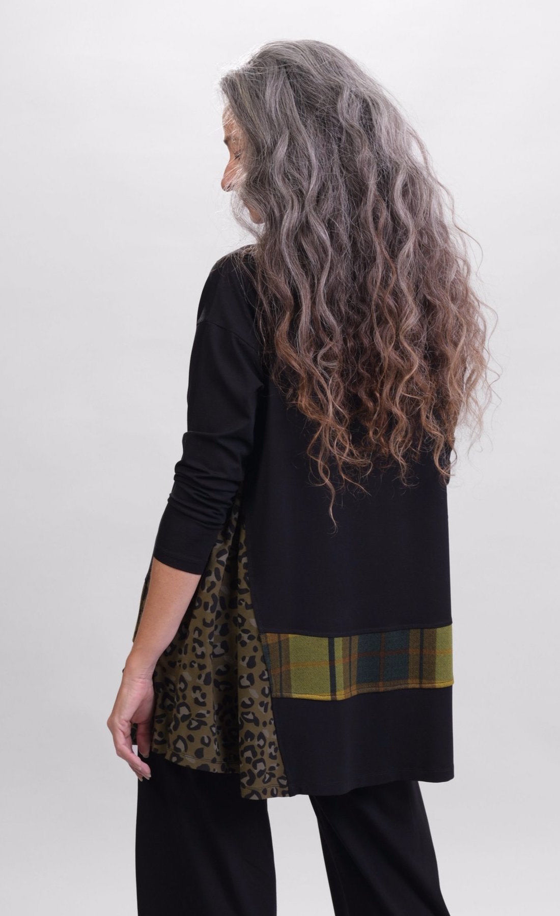 Back top half view of a woman wearing black pants and the alembika plaid asymmetry top. This top is black with a cheetah print side panel on the left side, long sleeves, and a horizontal green and yellow plaid panel running across the bottom of the top. The from hem is asymmetrical.