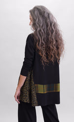 Load image into Gallery viewer, Back top half view of a woman wearing black pants and the alembika plaid asymmetry top. This top is black with a cheetah print side panel on the left side, long sleeves, and a horizontal green and yellow plaid panel running across the bottom of the top. The from hem is asymmetrical.
