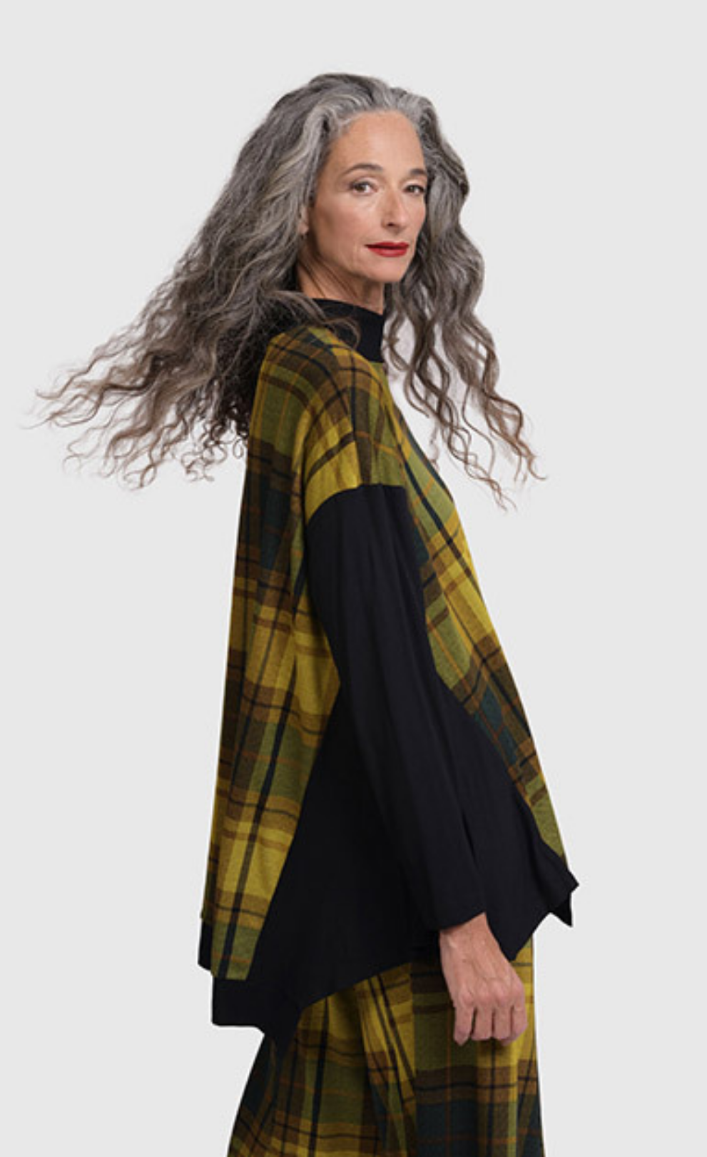 Right side top half view of a woman wearing black pants and the alembika plaid trapeze top. This top is a yellow and green plaid with solid black drop shoulder sleeves. The top has a funnel neck, side slits, and black detailing around the hem