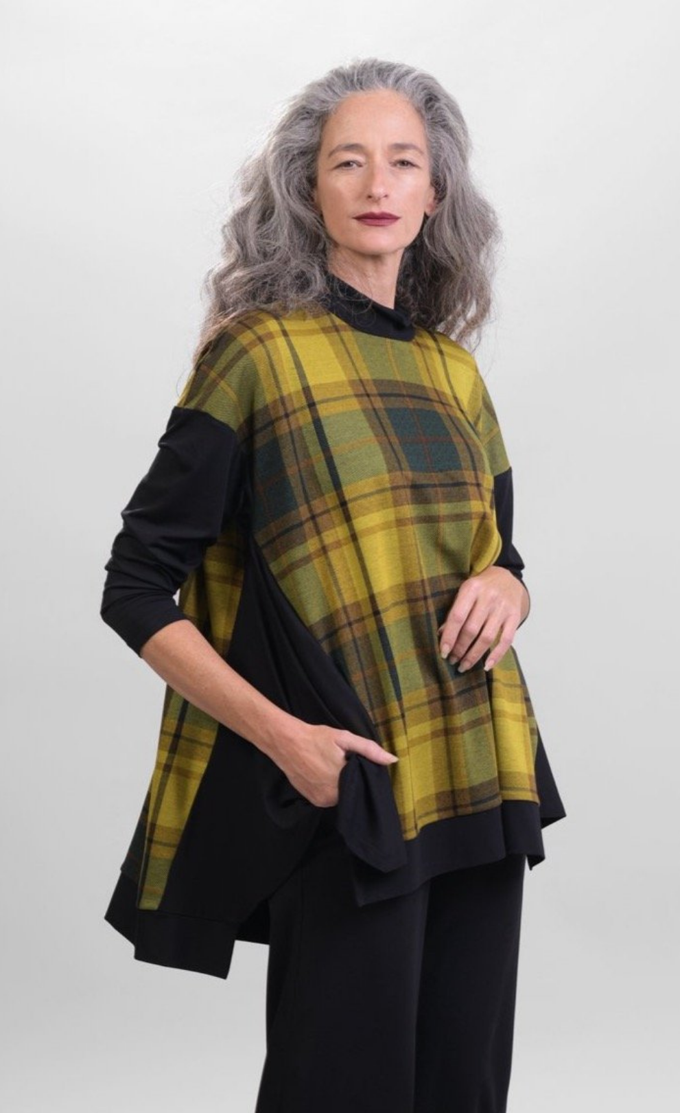 Front top half view of a woman wearing black pants and the alembika plaid trapeze top. This top is a yellow and green plaid with solid black drop shoulder sleeves. The top has a funnel neck, side slits, and black detailing around the hem