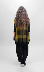 Load image into Gallery viewer, Back view of a woman wearing black pants and the alembika plaid trapeze top. This top is a yellow and green plaid with solid black drop shoulder long sleeves. The top has black detailing around the hem
