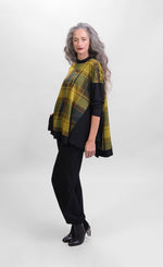 Load image into Gallery viewer, Left side full body view of a woman wearing black pants and the alembika plaid trapeze top. This top is a yellow and green plaid with solid black drop shoulder sleeves. The top has a funnel neck, side slits, and black detailing around the hem
