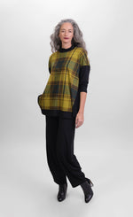 Load image into Gallery viewer, Front full body view of a woman wearing black pants and the alembika plaid trapeze top. This top is a yellow and green plaid with solid black drop shoulder sleeves. The top has a funnel neck, side slits, and black detailing around the hem
