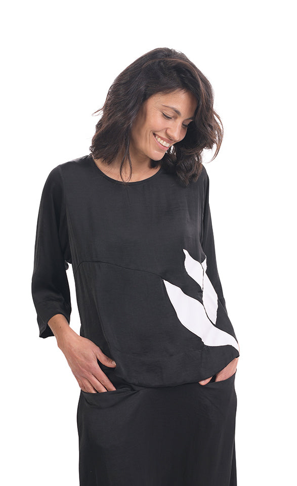 Front top half view of a woman wearing black pants and the alembika lotus top in black. This top has a round neck, 3/4 length sleeves, and a white lotus flower on the front left side.