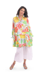 Load image into Gallery viewer, Front full body view of a woman wearing white wide pants and the alembika bloom getaway tunic shirt. This shirt has a bright green, pink, and yellow print, a button down front with a shirt collar, long sleeves with cuffs, and a gathered flounce hem.
