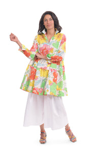 Front full body view of a woman wearing white wide pants and the alembika bloom getaway tunic shirt. This shirt has a bright green, pink, and yellow print, a button down front with a shirt collar, long sleeves with cuffs, and a gathered flounce hem.