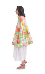 Load image into Gallery viewer, Left side full body view of a woman wearing white wide pants and the alembika bloom getaway tunic shirt. This shirt has a bright green, pink, and yellow print, a button down front with a shirt collar, long sleeves with cuffs, and a gathered flounce hem.
