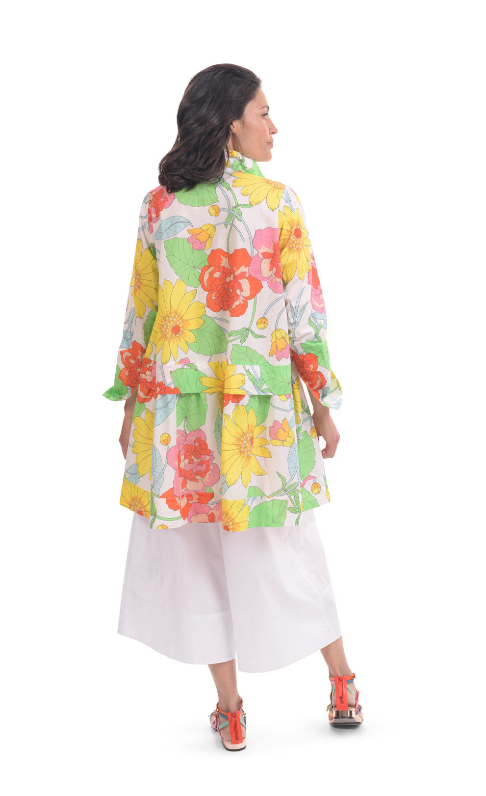 Back full body view of a woman wearing white wide pants and the alembika bloom getaway tunic shirt. This shirt has a bright green, pink, and yellow print, long sleeves with cuffs, and a gathered flounce hem.