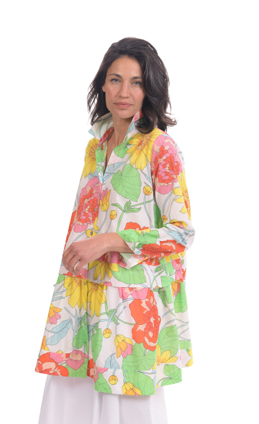 Front top half view of a woman wearing the alembika bloom getaway tunic shirt. This shirt has a bright green, pink, and yellow print, a button down front with a shirt collar, long sleeves with cuffs, and a gathered flounce hem.