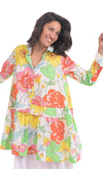 Load image into Gallery viewer, Front top half view of a woman wearing the alembika bloom getaway tunic shirt. This shirt has a bright green, pink, and yellow print, a button down front with a shirt collar, long sleeves with cuffs, and a gathered flounce hem.
