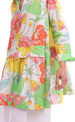 Load image into Gallery viewer, Front close up view of a woman wearing the alembika bloom getaway tunic shirt. This shirt has a bright green, pink, and yellow print, a button down front with a shirt collar, long sleeves with cuffs, and a gathered flounce hem.
