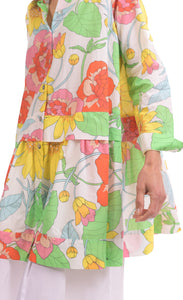 Front close up view of a woman wearing the alembika bloom getaway tunic shirt. This shirt has a bright green, pink, and yellow print, a button down front with a shirt collar, long sleeves with cuffs, and a gathered flounce hem.