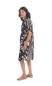 Left side full body view of a woman wearing the alembika Speckled Shayna Shirtdress. This shirtdress has a button down front, short sleeves, and a black and white tie-dye print on the front with a black and white striping on the back.