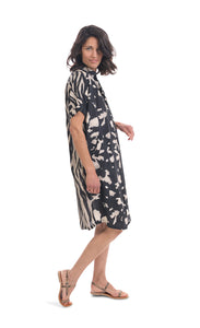 Right side full body view of a woman wearing the alembika Speckled Shayna Shirtdress. This shirtdress has a button down front, short sleeves, and a black and white tie-dye print on the front with a black and white wavy striping print on the back