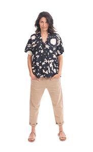 Front full body view of a woman wearing khaki pants and the alembika speckle mandala shirt. This shirt has a black and white mandala tie dye print, a button down front, a shirt collar, a boxy silhouette, and short sleeves.