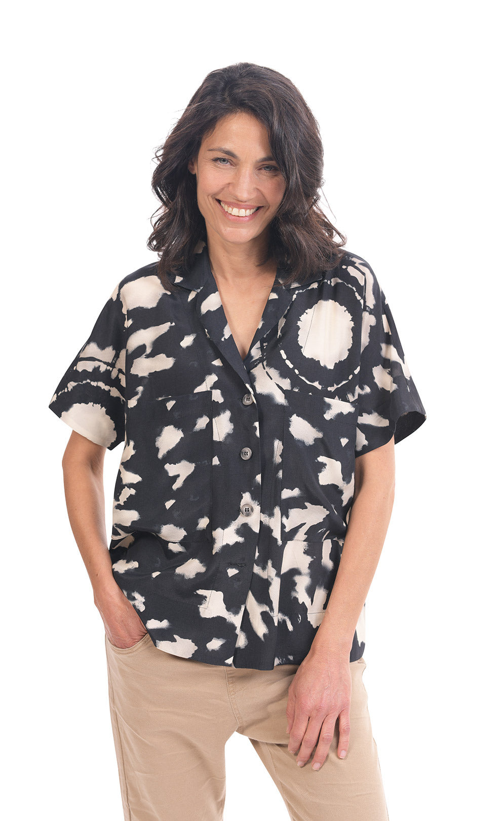 Front top half view of a woman wearing khaki pants and the alembika speckle mandala shirt. This shirt has a black and white mandala tie dye print, a button down front, a shirt collar, a boxy silhouette, and short sleeves.