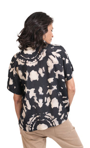 Back top half view of a woman wearing khaki pants and the alembika speckle mandala shirt. This shirt has a black and white mandala tie dye print, a boxy silhouette, and short sleeves.