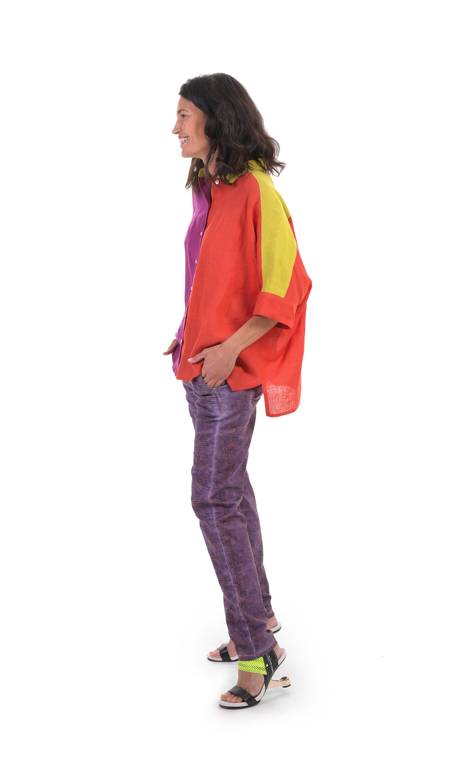 Left side full body view of a woman wearing the alembika colorblock linen shirt. This shirt is red on the left side, purple on the right side and yellow on the sleeves and shoulders. The shirt has a button up front, a shirt collar, and elbow length sleeves. On the bottom, the model is wearing purple pants.