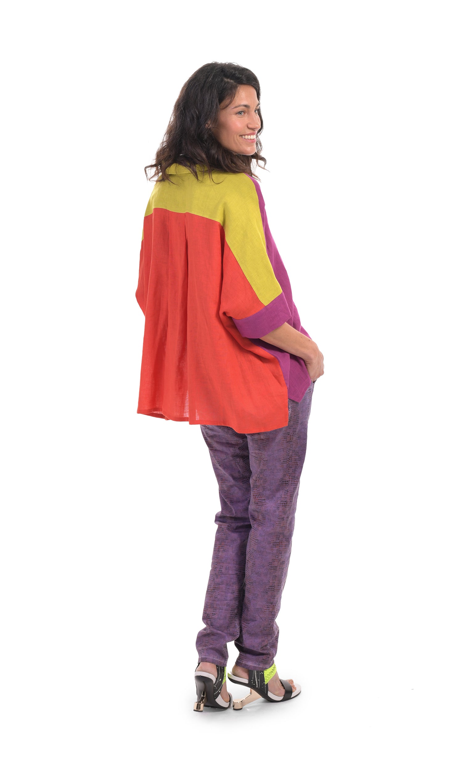 Back full body view of a woman wearing the alembika colorblock linen shirt. This shirt is red on the back, purple on the right side and yellow on shoulder and sleeve sides. The shirt has a button up front, a shirt collar, and elbow length sleeves. On the bottom, the model is wearing purple pants.