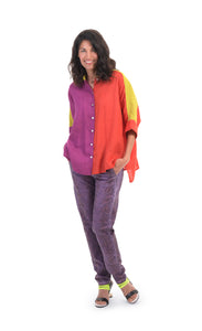 Front full body view of a woman wearing the alembika colorblock linen shirt. This shirt is red on the left side, purple on the right side and yellow on the shoulders and sleeves. The shirt has a button up front, a shirt collar, and elbow length sleeves. On the bottom, the model is wearing purple pants.