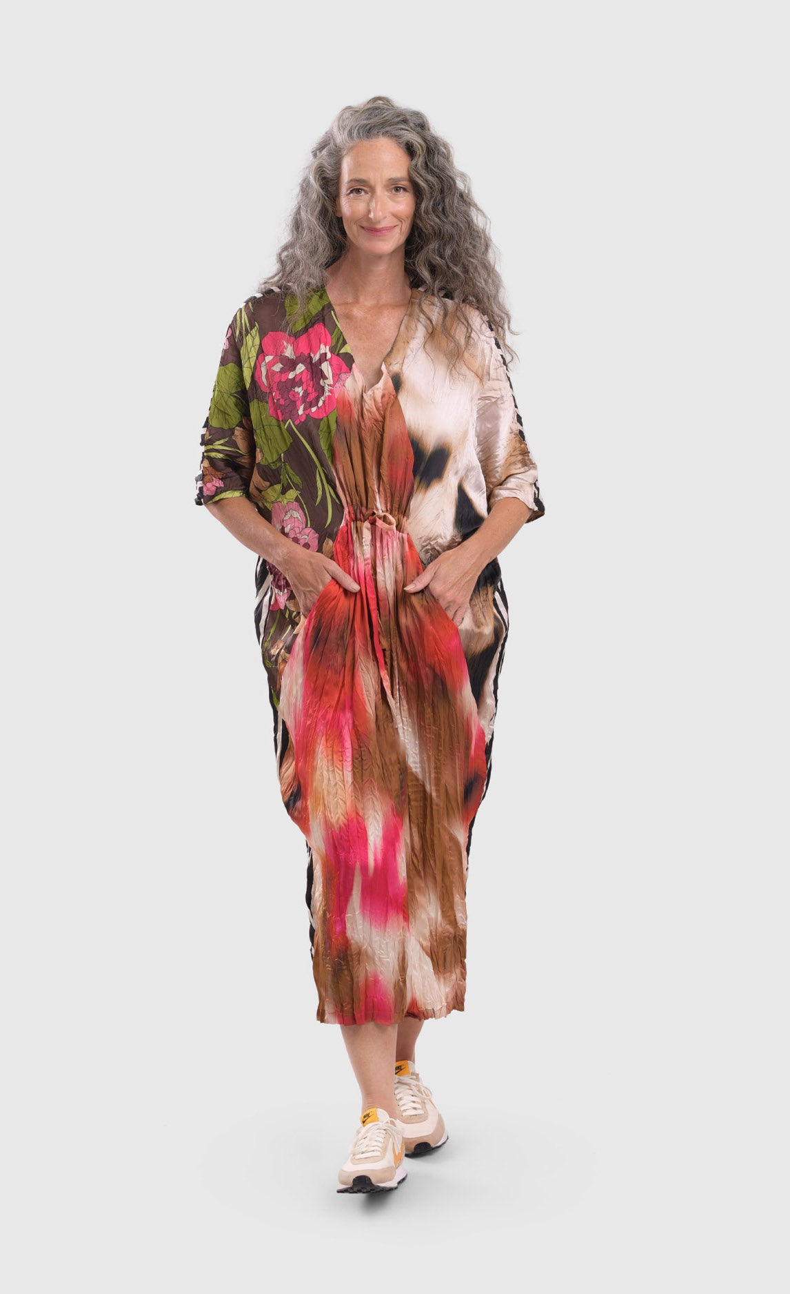 Front full body view of a woman wearing the alembika skye caftan butterfly dress. This dress has pink flowers over a brown backdrop on the right side, a pink and brown tie dye print in the center, and a beige and brown tie dye print on the left side.