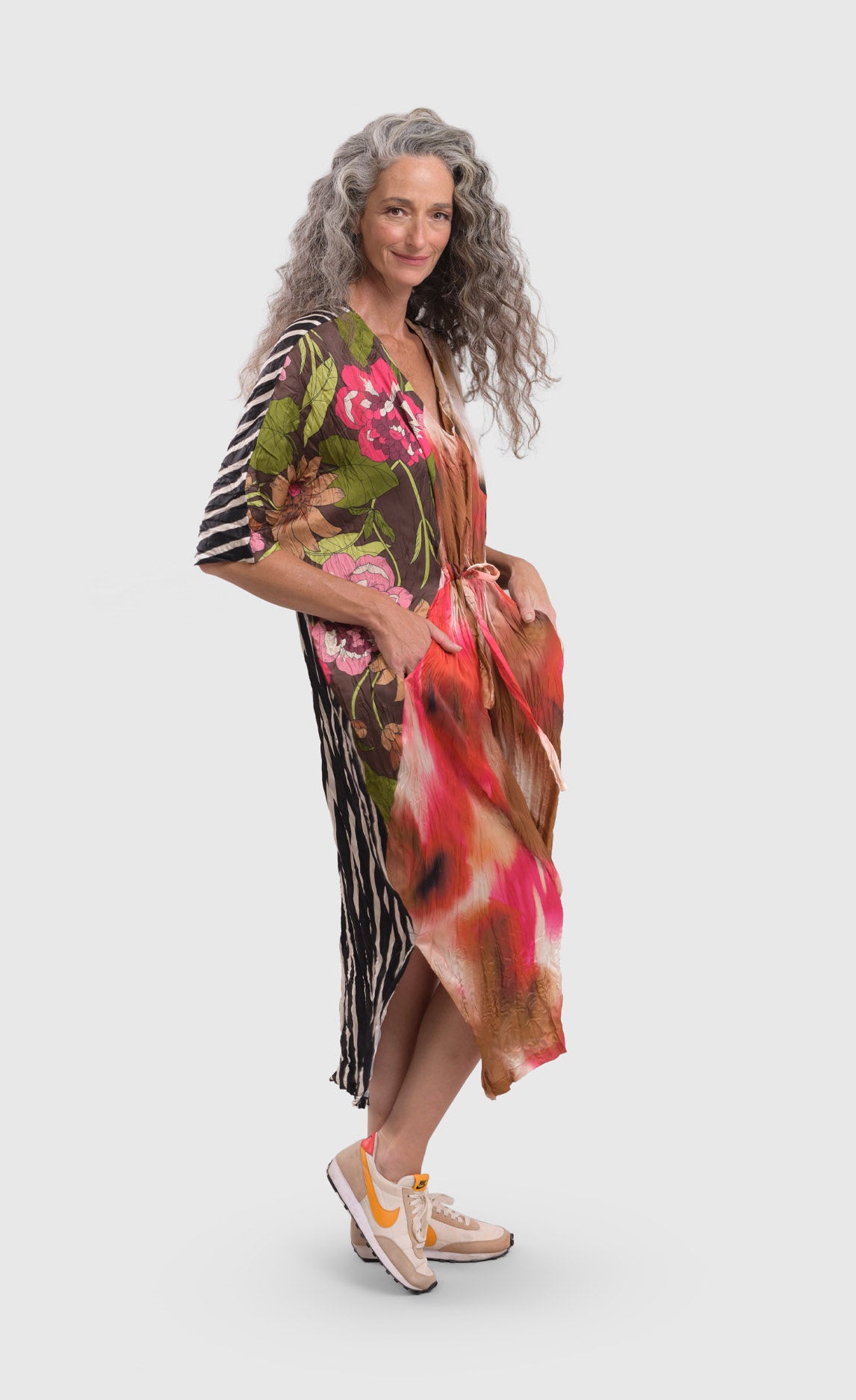 Front right side full body view of a woman wearing the alembika skye caftan butterfly dress. This dress has pink flowers over a brown backdrop on the right side, a pink and brown tie dye print in the center, a beige and brown tie dye print on the left side, and a black and white striped print on the back.