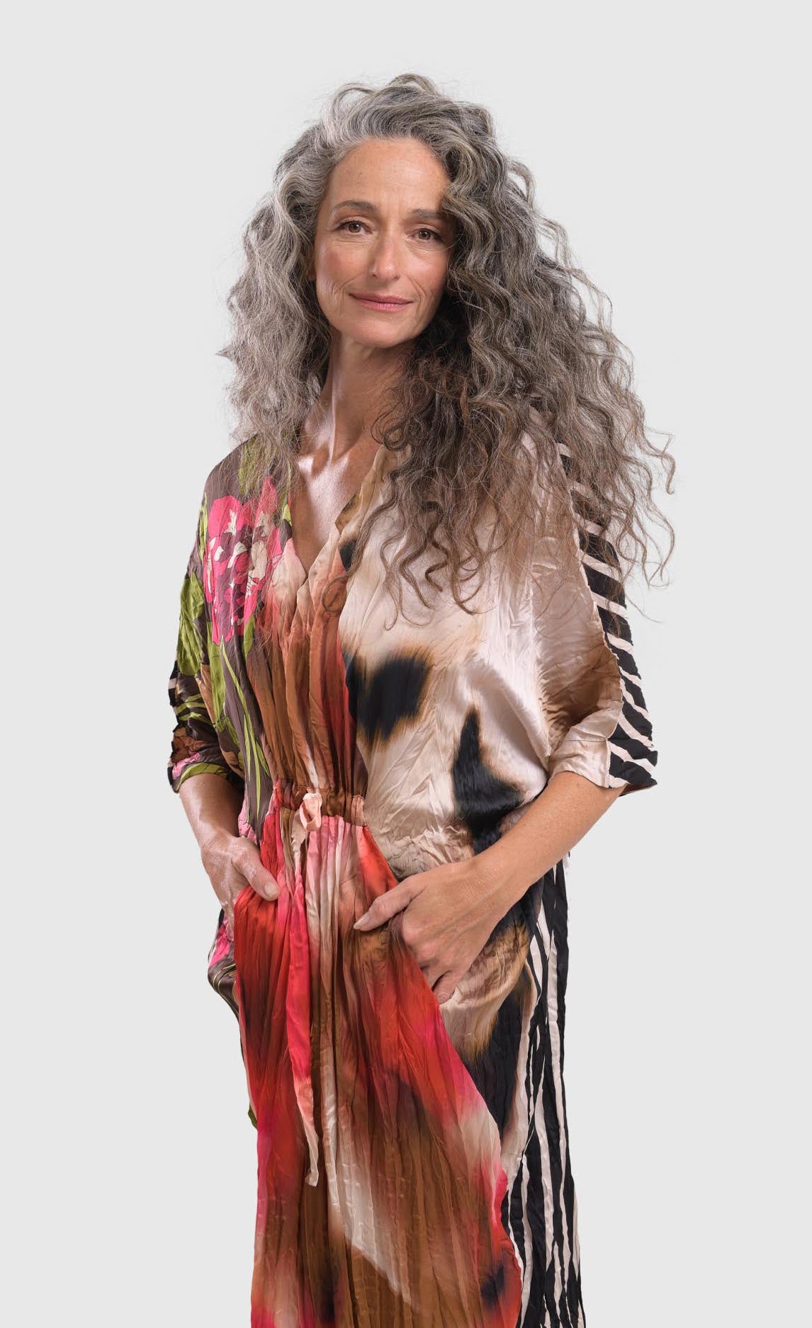 Front left side top half view of a woman wearing the alembika skye caftan butterfly dress. This dress has pink flowers over a brown backdrop on the right side, a pink and brown tie dye print in the center, a beige and brown tie dye print on the left side, and a black and white striped print on the back.